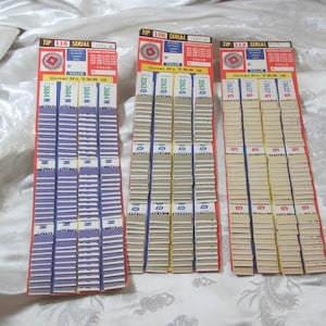 Può includere: Tre tabelloni rettangolari con intestazioni rosse, ciascuno con un logo circolare, tabelle numeriche e il testo Order No. T.E.R. 18. I tabelloni contengono file di biglietti a strappo nei colori viola, bianco e marrone chiaro con numeri di serie.