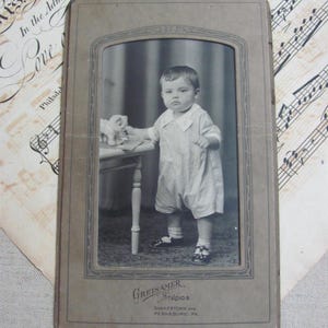 May include: Vintage black and white studio portrait of a baby in a romper standing next to a small table with a toy. The photo is in a decorative frame with the studio name "GREISAMER STUDIOS" and location details.