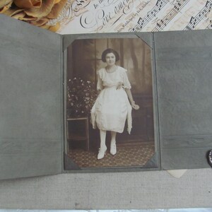 May include: Vintage sepia-toned photograph of a woman in a white dress, posed inside a gray photo album. The album is open, revealing the portrait. The woman is wearing a dress with short sleeves and white shoes. The background includes floral arrangements and sheet music.