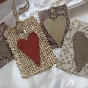May include: Four fabric gift tags with heart shapes. The tags are made of burlap, tweed, and patterned fabric. The hearts are made of brown and red suede.