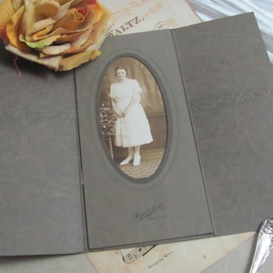 May include: A vintage sepia-toned photograph of a woman in a white dress, set within a gray, tri-fold photo album. A large, fabric rose in shades of yellow and orange rests above the album. Additional vintage ephemera is visible.
