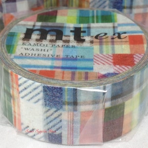 Puede incluir: Un rollo de cinta adhesiva mt ex Kamoi Paper "Washi". La cinta presenta un diseño de patchwork con cuadrados de cuadros en varios colores, incluyendo rojo, azul, verde y amarillo. La cinta está envuelta en plástico transparente.