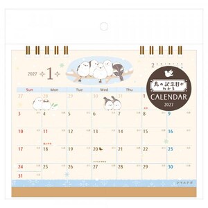 May include: A 2027 desk calendar featuring illustrations of small white birds on branches. The page shows January with a grid of dates, snowflake accents, and a circular dark brown badge with the text 鳥の記念日がわかる CALENDAR 2027. The calendar is bound with a gold-coloured metal spiral at the top.