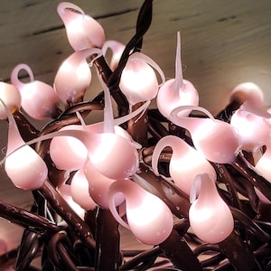 May include: A string of pink fairy lights with white bulbs. The lights are in the shape of teardrops and are strung on a brown wire.