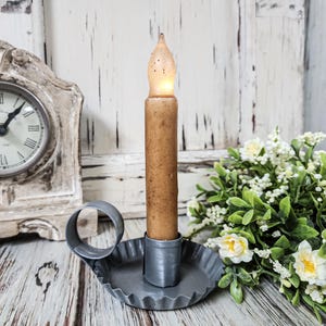 May include: A battery-operated candle with a flickering amber LED flame sits in a gray metal holder. The candle has a textured, brown wax appearance. The holder has a scalloped edge and a handle. A vintage clock and floral arrangement are in the background.