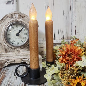 May include: Two battery-operated, brown taper candles with flickering amber LED lights. The candles are in black metal holders. A vintage-style clock and fall floral arrangement are in the background.