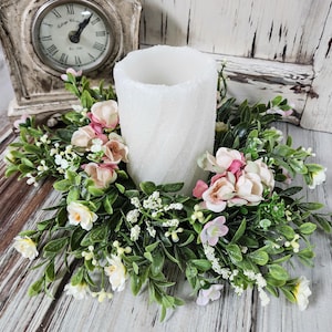 May include: A white pillar candle surrounded by a floral wreath with pink and white flowers and green leaves. The candle sits on a wooden surface, with a vintage clock in the background.