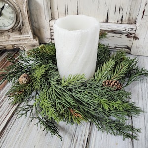 May include: A white, textured pillar candle sits in a green evergreen wreath with pinecones. The candle has a swirling pattern and is surrounded by realistic-looking greenery. The arrangement is on a rustic, white-washed wooden surface.