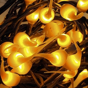May include: A string of decorative lights featuring numerous small, translucent, amber-colored bulb covers shaped like teardrops or flames. The bulbs are clustered together and attached to dark, intertwined electrical wires, emitting a warm glow.