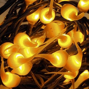 May include: A string of decorative lights featuring numerous small, translucent, amber-colored bulb covers shaped like teardrops or flames. The bulbs are clustered together and attached to dark, intertwined electrical wires, emitting a warm glow.
