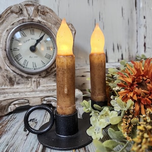 May include: Two battery-operated, brown, speckled taper candles with flickering amber flames. The candles are in a black metal candle holder with a ring handle. A decorative clock and orange flowers are in the background.