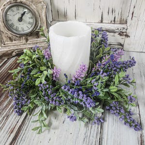 May include: A white pillar candle surrounded by a wreath of green leaves and purple and lavender flowers. The candle sits on a distressed wooden surface. A vintage clock is in the background.