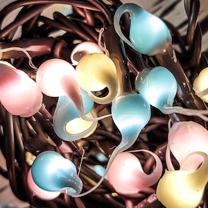 May include: Close-up of a string of decorative lights with teardrop-shaped bulbs. The bulbs are in various pastel colors, including pink, yellow, and blue. The lights are strung on dark brown wires, creating a warm and inviting glow.