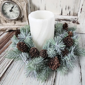 May include: A white pillar candle sits in a wreath of green and blue-green pine needles and brown pine cones. The candle and wreath are on a distressed white wooden surface. A vintage clock is in the background.