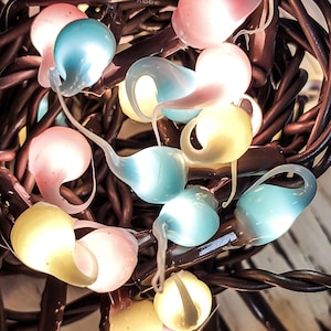 May include: Close-up of a string of decorative lights with teardrop-shaped bulbs in pastel colors. The bulbs are pink, blue, and yellow, and are strung on dark brown wires. The lights are lit, creating a soft glow.