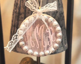 May include: A circular ornament featuring a light pink marbled background with a white script monogram of the letters MMR. The edge is decorated with a border of white faux pearls and a white lace ribbon tied in a bow at the top.