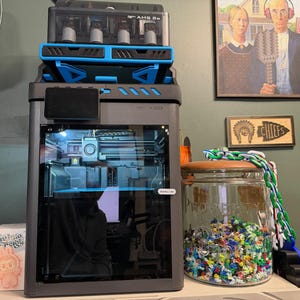 May include: A dark gray 3D printer with a blue and black AMS unit on top, featuring a glass front door and a small digital screen. Next to it sits a large clear glass jar filled with colorful plastic scraps and a green and white chain.