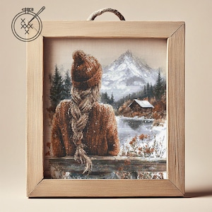 May include: A cross-stitch embroidery framed in a light brown wood frame. The embroidery depicts a woman with long braided hair wearing a brown hat and a brown sweater, sitting on a bench overlooking a snowy lake and a cabin in the distance. A mountain with a snowy peak is in the background.