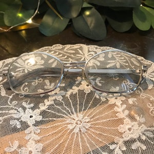 May include: Silver-toned oval-shaped eyeglasses with a thin metal frame. The glasses are resting on a white lace doily.