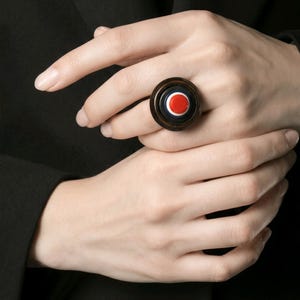 May include: A large, round ring with a brown metal band. The ring's face features a design with a red circle in the center, surrounded by white and navy blue concentric circles. The ring is worn on a finger.
