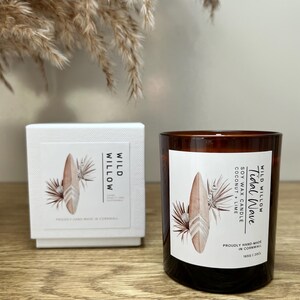 May include: A brown glass candle with a white label featuring a surfboard illustration and the text "Tidal Wave". The candle is next to a white box with matching design elements. The candle is soy wax with coconut and lime scent.