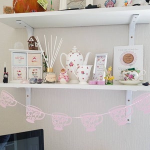 May include: A white shelf with a pink teacup garland hanging from it. The shelf is decorated with various items, including a small wooden house, a teapot, a vase with flowers, and a framed picture. The garland is made of pink paper cutouts of teacups.