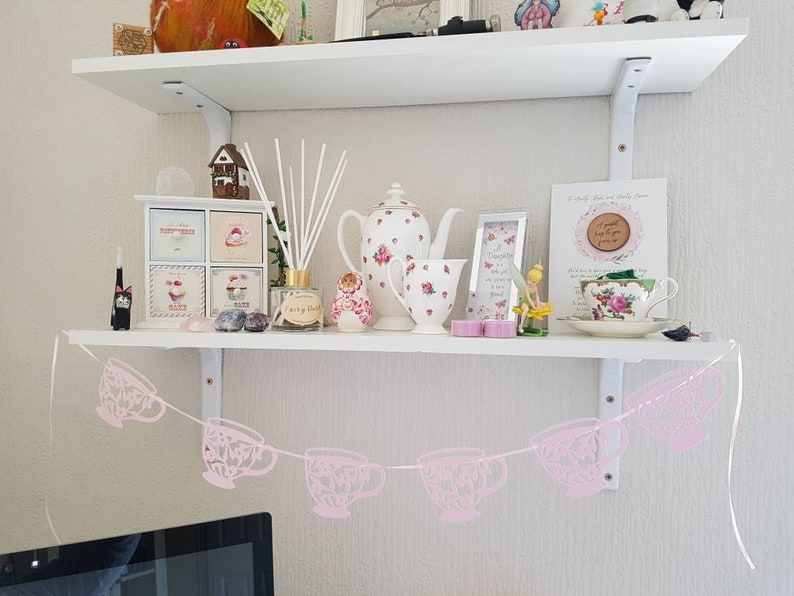 May include: A white shelf with a pink teacup garland hanging from it. The shelf is decorated with various items, including a small wooden house, a teapot, a vase with flowers, and a framed picture. The garland is made of pink paper cutouts of teacups.