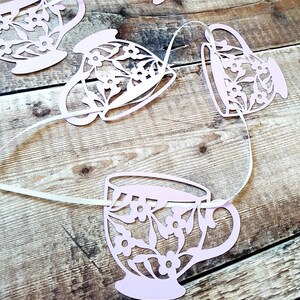May include: A pink paper garland with a string of teacups, each with a floral design. The teacups are cut out and strung together with white ribbon.