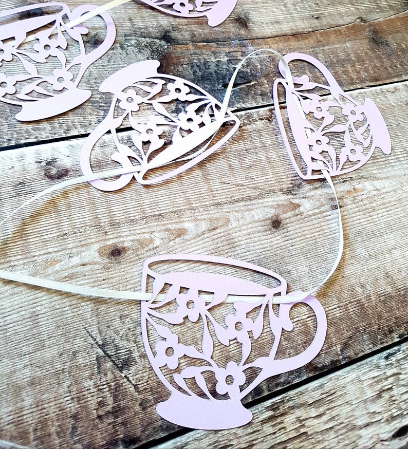 May include: A pink paper garland with a string of teacups, each with a floral design. The teacups are cut out and strung together with white ribbon.
