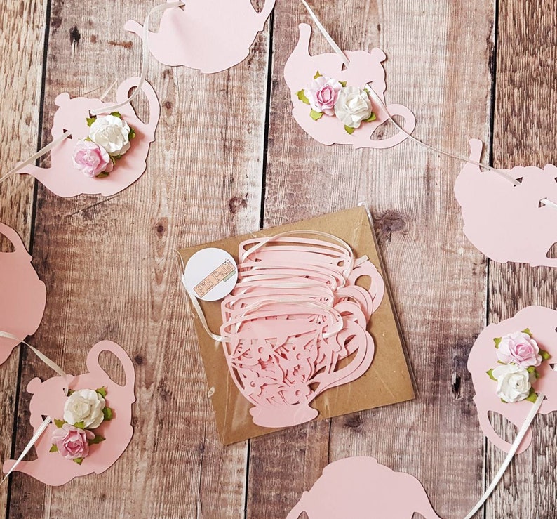 May include: Pink paper teacup and teapot garland with white flowers. The garland is strung together with white ribbon. The teacups and teapots are cut out of paper and have a delicate design.
