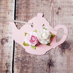 May include: Pink paper teapot with two white and pink fabric flowers attached to it. The teapot is strung with white ribbon.