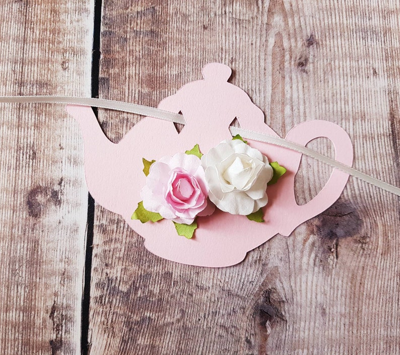 May include: Pink paper teapot with two white and pink fabric flowers attached to it. The teapot is strung with white ribbon.