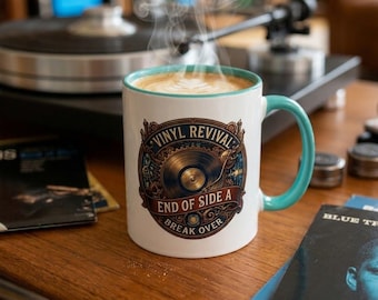 May include: A white ceramic mug with a teal rim and handle, featuring a graphic of a vinyl record and the text "VINYL REVIVAL END OF SIDE A BREAK OVER." The mug contains a steaming hot beverage with latte art, positioned on a wooden surface.
