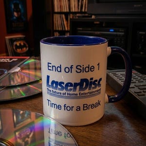 Puede incluir: Una taza de cerámica blanca con interior y asa de color azul oscuro. El frente presenta texto azul que dice End of Side 1, LaserDisc, The future of Home Entertainment y Time for a Break, con líneas horizontales junto al logotipo.