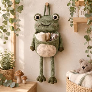 May include: A green crocheted frog wall hanging with a large storage pocket on its belly. The frog features large black eyes, a stitched smile, and long dangling legs. Inside the pocket are a folded white cloth, a wooden brush, and a small toy.