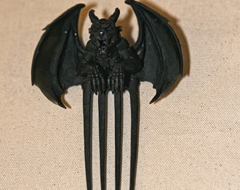 May include: A matte black hair comb featuring a sculpted gargoyle figure at the top. The gargoyle has large, spread wings, pointed ears, and a snarling expression. The comb has four long, slender, pointed tines extending downward from the base.