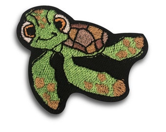 May include: Embroidered patch featuring a cartoon sea turtle. The turtle has a green body with brown and tan accents on its shell. The patch is outlined in black and has a friendly expression.