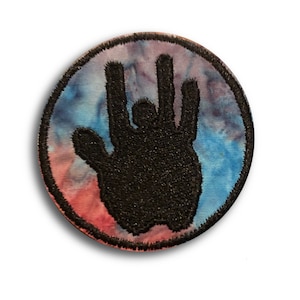 May include: A circular patch with a black hand gesture design set against a tie-dye effect of blue, pink, and purple. The patch has a black border and is likely an iron-on or sew-on patch.