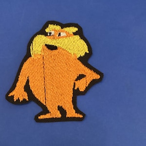May include: Embroidered patch of a cartoon character with orange fur, yellow hair, and a black border. The patch is set against a blue backdrop. The character has a mustache and is smiling.