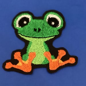 Pode incluir: Patch bordado com um desenho de um sapo de desenho animado. O sapo é verde com olhos grandes com bordas brancas e patas laranja. O patch tem uma borda preta e está sobre um fundo azul. Ideal para roupas ou acessórios.