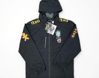 Peut inclure: Un coupe-vent noir à capuche avec le texte jaune Itaú sur les épaules et Vivo sur la manche. La poitrine affiche l'emblème de l'équipe nationale de football du Brésil et un petit motif orange. Une étiquette blanche est attachée à la fermeture.