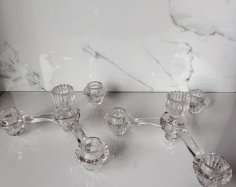 May include: Two clear glass candelabras, each featuring a three-armed design with ribbed cylindrical candle holders at the ends of each arm. The glass is transparent and smooth, with a faceted texture on the base of each candle cup.