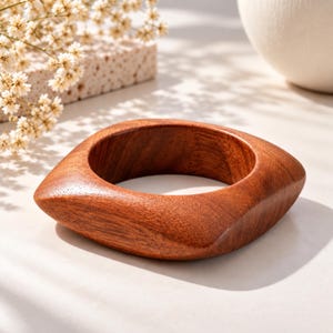 May include: A smooth, polished brown wooden bangle bracelet with a rounded square shape. The bracelet features a rich, warm wood grain pattern and a thick, solid construction, showcasing a natural finish with visible wood texture throughout the piece.