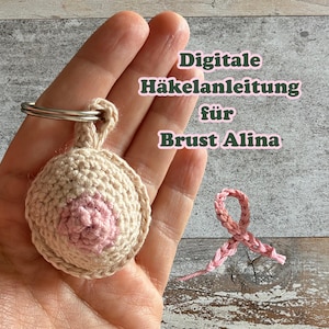 Könnte beinhalten: Ein beigefarbener und hellrosa gehäkelter Schlüsselanhänger in Brustform mit einem Metallring und einer separaten kleinen rosa gehäkelten Schleife. Das Bild enthält den Text: Digitale Häkelanleitung für Brust Alina.