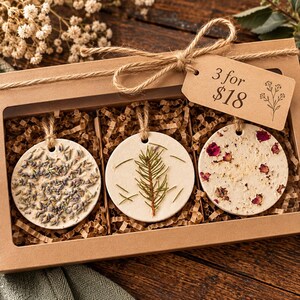 May include: A brown cardboard gift box containing three round, off-white clay ornaments hanging from jute twine. The ornaments feature embedded dried lavender, a pine sprig, and dried rose petals. A tag attached to the box reads 3 for $18.