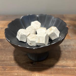 May include: A black pedestal bowl with a scalloped edge contains several small, light gray, hexagonal-shaped objects. The bowl has a matte finish and a wide, shallow basin, while the gray items appear solid with a textured, stone-like surface.