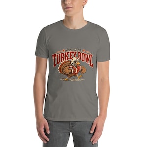 May include: A grey short-sleeve t-shirt featuring a graphic of a turkey wearing a football helmet and jersey, holding a football. The text above the turkey reads ANNUAL TURKEY BOWL in red letters with orange accents and two small stars.