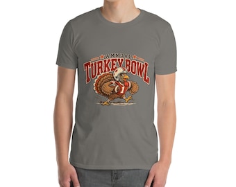 May include: A grey short-sleeve t-shirt featuring a graphic of a turkey wearing a football helmet and jersey, holding a football. The text above the turkey reads ANNUAL TURKEY BOWL in red letters with orange accents and two small stars.