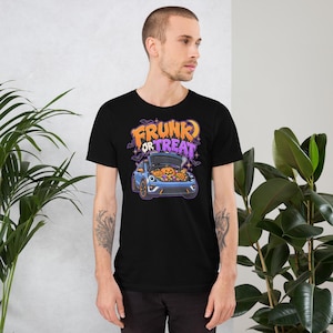 May include: A black short-sleeve t-shirt featuring a graphic of a blue car with its front trunk open, filled with orange jack-o'-lantern pumpkins. Above the car, the text reads FRUNK OR TREAT in orange and purple stylized lettering.