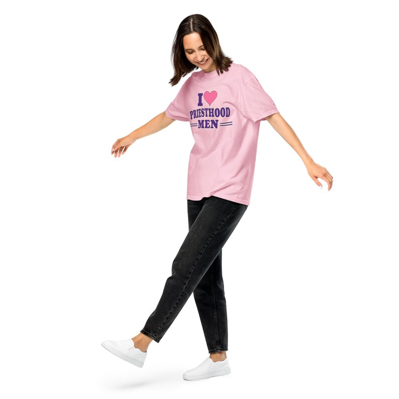 May include: A light pink short-sleeve t-shirt featuring a graphic print with a pink heart icon above the purple text that reads I HEART PRIESTHOOD MEN with two horizontal lines below the word MEN, paired with black denim jeans and white sneakers.